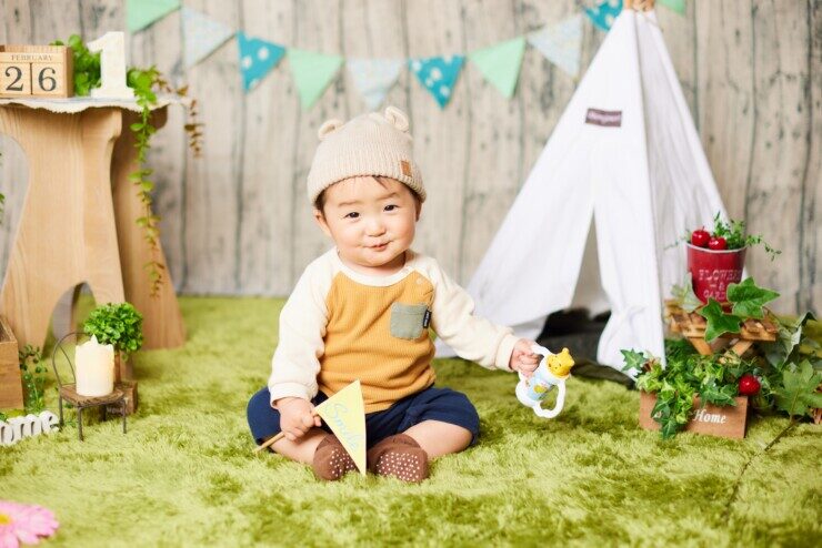 赤ちゃんが旗を持ち、緑色の絨毯の上でポーズを取り、首をかしげて、ほほ笑んでいるFamm無料撮影会の光景